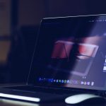 Open laptop on a desk with a dark screen, accompanied by a mouse and a cup in a cozy, dimly lit setting.