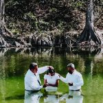 Baptism ceremony in a serene river surrounded by large trees, with three people wearing white shirts.