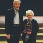 Elderly couple celebrates award on stage, holding plaque. Man on left, woman on right smiling.