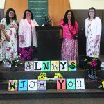 Group of women in floral dresses standing by a podium with colorful signs reading ALWAYS WITH YOU.