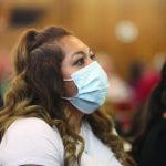 Woman wearing a mask listens attentively in a room with blurred background.