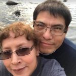 Couple taking a selfie by the river, smiling and wearing glasses, with rocks in the water background.