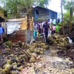People walking along a rocky path beside makeshift houses in a rural area.