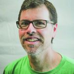 Smiling person wearing glasses and a green shirt against a plain background.