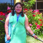 Woman in green dress standing by garden with red flowers, smiling at camera.