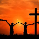 Silhouetted family holding hands beside a cross at sunset, symbolizing unity and faith.