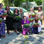 Group of women smiling and holding colorful flower pots outdoors on a sunny day.