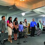 Group of people singing on stage in a decorated room, with musical instruments in the background.
