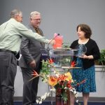 Three people at a podium exchanging a book and an award, surrounded by colorful flowers and greenery.