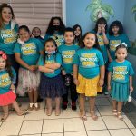 Children and a teacher in matching teal I Am Priceless to God shirts in a colorful room with palm tree decor.
