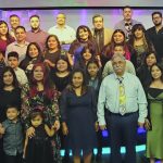 Group photo of diverse people posing together indoors on a brightly lit stage with decorative background.