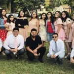 Group of people gathered outdoors on grass, smiling, dressed in formal and casual attire, with trees and houses in background.