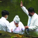 Baptism ceremony in river, person being baptized, two others assisting, wearing masks and white attire.