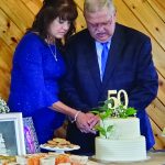 Couple cutting 50th anniversary cake at a celebration, surrounded by snacks and a framed photo with smiles.