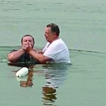 Two people performing a baptism in a calm lake, one submerged, guided by the other in a white shirt.
