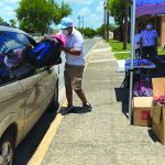 Man loading backpacks into a van at outdoor community event with tents and volunteers.