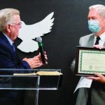 Two men at an award ceremony, one holding a framed certificate, microphone on stand nearby.