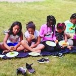 Children having a picnic on a grassy field, sitting on a blanket with plates of food, and enjoying a sunny day outdoors.