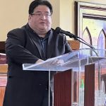 A person speaking at a podium with a microphone in a well-lit room, wearing a dark suit.
