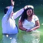 Person in water performing a baptism ceremony with woman wearing a floral crown, under bright sunlight.