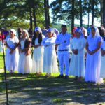 Outdoor ceremony with people in white attire gathered by a lake under trees, listening to a speaker at a podium.
