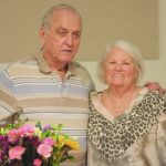 Elderly couple smiling together indoors, standing beside a table with colorful flowers in vase.