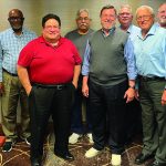 A group of nine diverse businessmen smiling and posing together in a conference room setting.