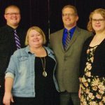 A group of four people smiling, posing together at an indoor event, dressed in formal and casual attire.