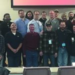 Group of men gathered indoors for a meeting or seminar, posing for a photo, with a screen in the background.