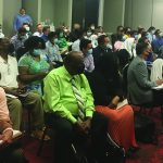 Audience seated in a conference room, wearing masks, attentively listening to a speaker.