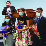 Three people holding bouquets with a speaker at a podium, all wearing masks, in an indoor setting.