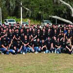 Group of people in matching black shirts posing outdoors on grass with trees in background.