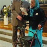 Elderly woman with a walker speaking at an event, assisted by a man wearing a suit and mask.