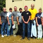 Group of people standing in front of a shed during daytime, wearing casual clothing and smiling.