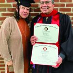 Graduate proudly holding two diplomas with a woman beside, both smiling in front of a brick wall.