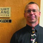 Man standing by office door labeled Pastor in formal attire, smiling.
