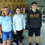 Three people smiling at a shoe donation event in a warehouse setting, surrounded by sorted footwear.