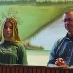 Two people standing by a serene landscape painting indoors.