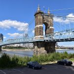 Historic suspension bridge with city skyline in background on sunny day, viewed from riverside parking area.