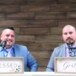 Two men in suits speaking at a wooden backdrop, signs with Blessed and Grateful in front of them.