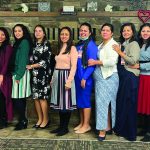 A diverse group of women standing together, smiling, in a warmly decorated room with heart and floral decor.