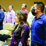 People standing in a conference room, wearing formal attire, some with masks, attending an event.
