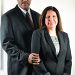 Formal portrait of a business couple in black suits, standing together against a white background.