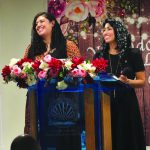 Two women smiling and speaking at a podium adorned with floral arrangements during a conference.