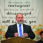 Man speaking at podium with encouraging banner behind. Message: Be strong and courageous.