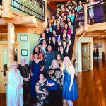 Group of women gathered on a staircase indoors, dressed in formal attire, smiling and posing for a photo.