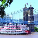 Paddle steamboat cruising under bridge in an urban river scene with clear blue sky and city skyline.
