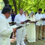 Group in white outfits reading from books outdoors in a wooded area, participating in a spiritual gathering or ceremony.