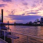River scene with paddleboat, sunset sky, and city skyline in the background, viewed from a deck with a covered table.