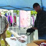 Outdoor market scene with people under tents, displaying clothes and serving snacks.